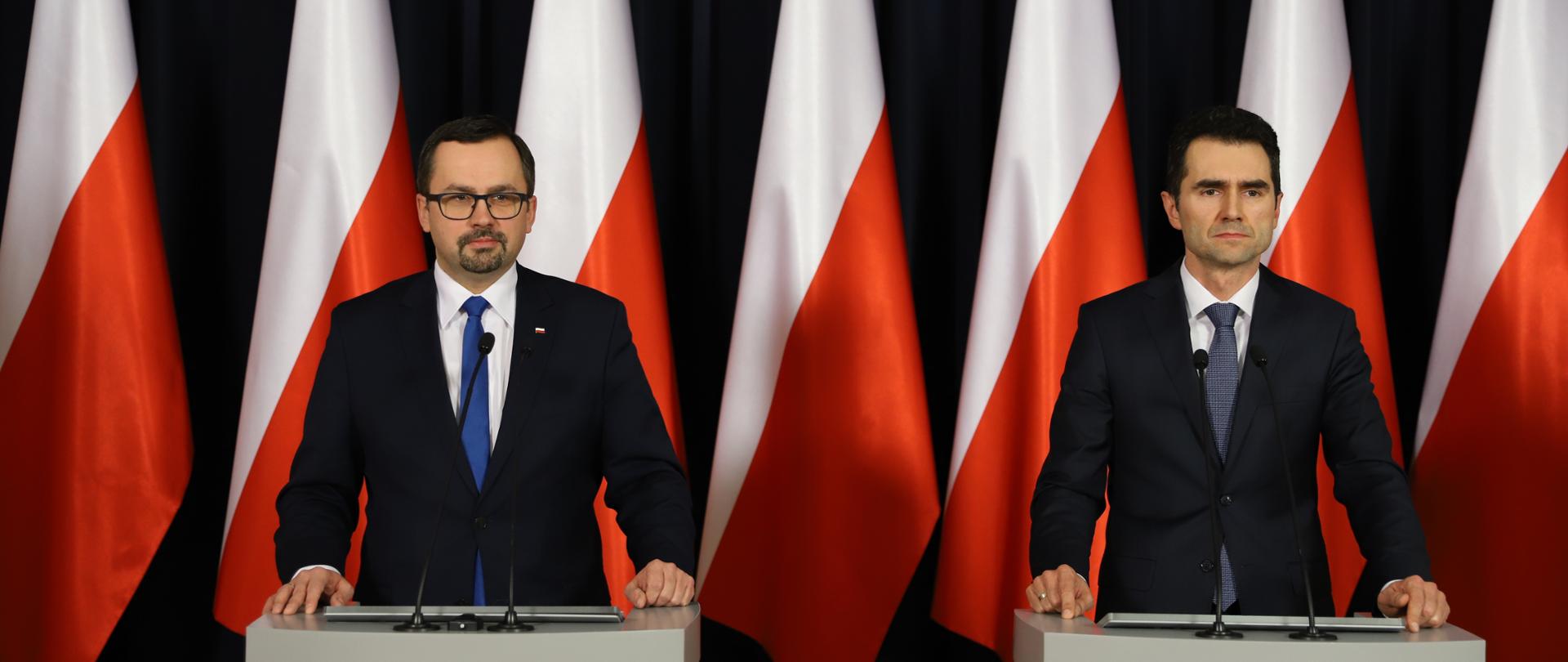 Na zdjęciu widać dwóch panów, którzy stoją przy mównicach. W tle widać biało - czerwone flagi