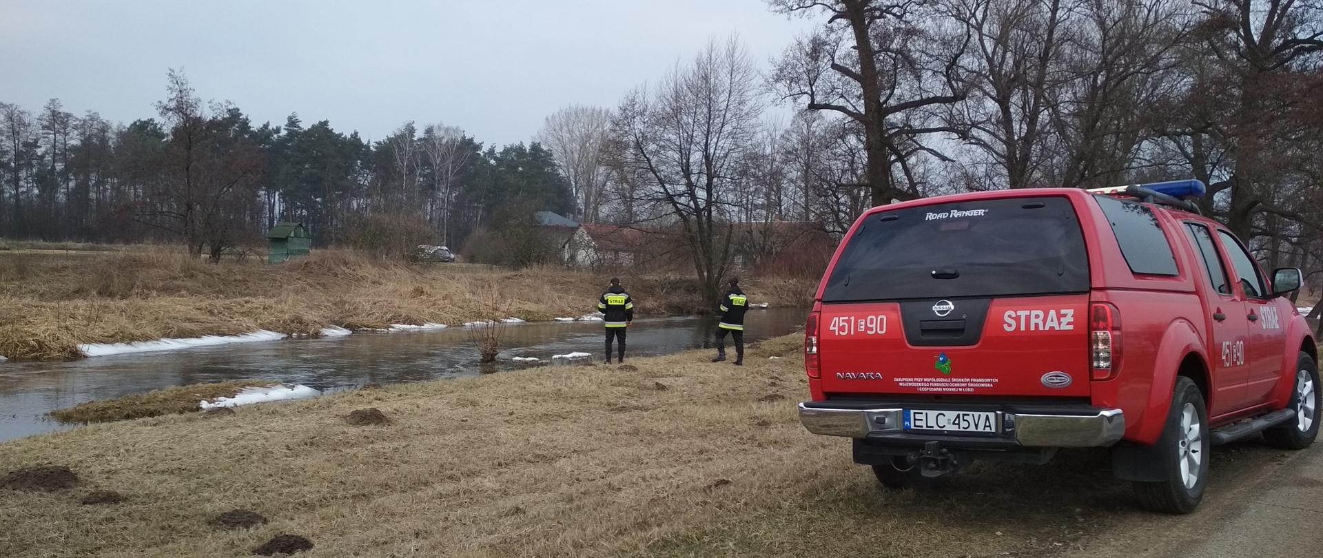 Na zdjęciu widać dwóch strażaków, którzy patrzą na rzekę, w tle rzeka , lasek, zabudowania. Na pierwszym planie po prawej terenowy samochód strażacki.