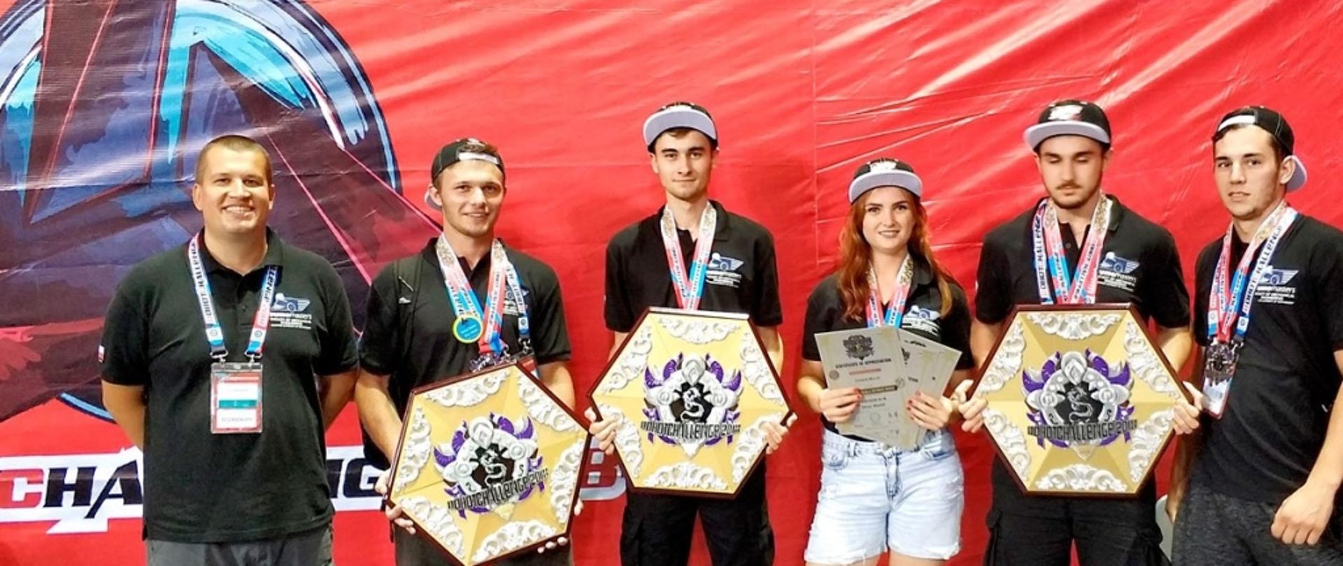 Na tle czerwonej ściany stoja studenci trzymający plaketki z medalami