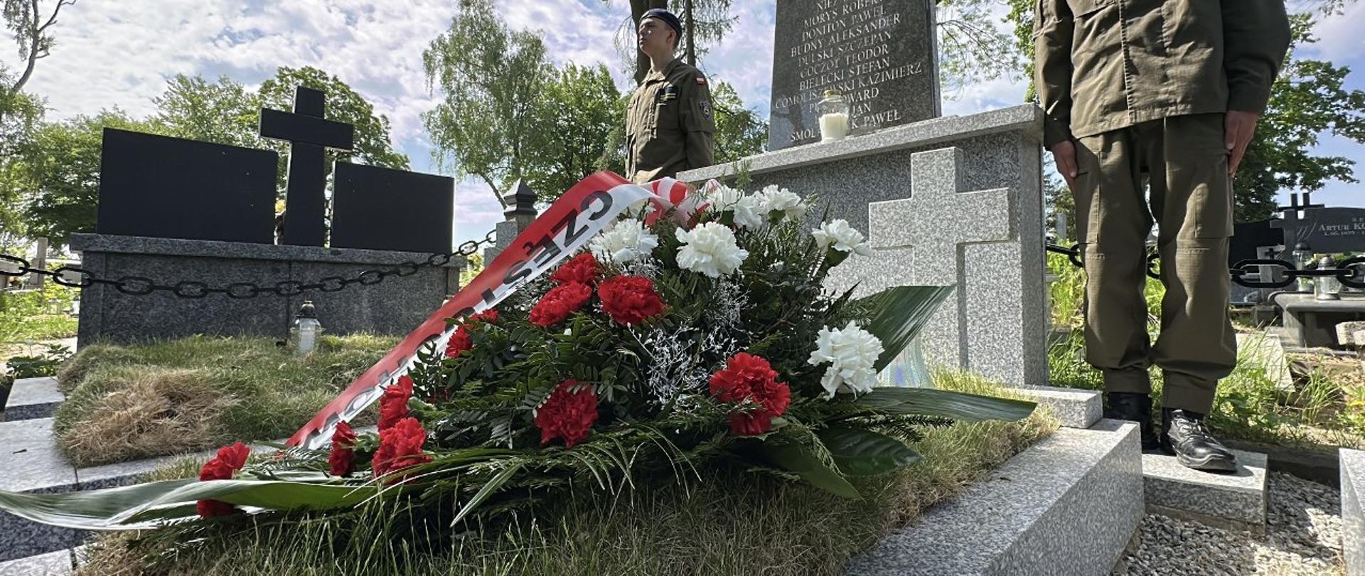 Grób na którym leży wieniec złożony z biało-czerwonych róż w głębi widoczny dwóch żołnierzy pełniących wartę przy pomniku