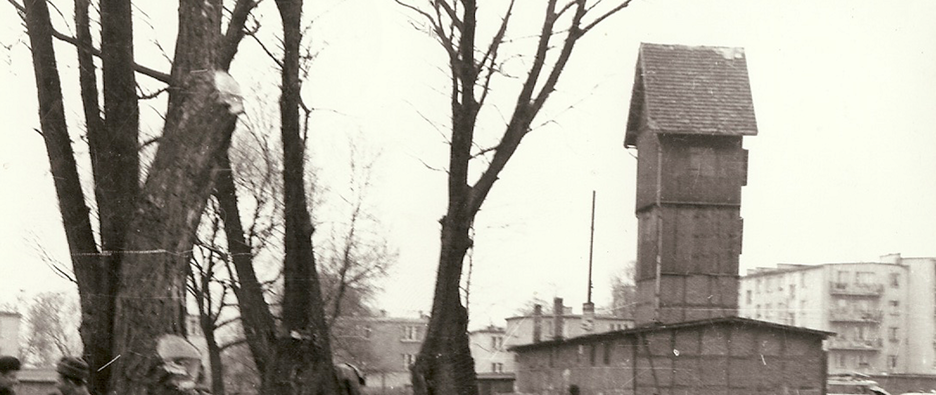 Na zdjęciu znajduje się budynek poniemieckiej strażnicy. Budynek widoczny w górnym prawym rogu zdjęcia. Obiekt to jednokondygnacyjny budynek z dwuspadzistym dachem. Po prawej stronie strażnicy widać wieżę pożarniczą. Po lewej stronie zdjęcia widoczne drzewa a za nimi rzeka iławka. Pomiędzy drzewami widać niewyraźne postacie pracujących ludzi. Zdjęcie czarno-białe.