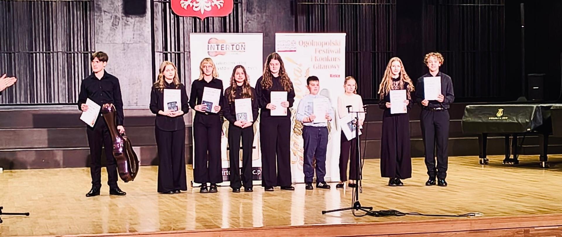 Kolorowe zdjęcie, na którym można zobaczyć 9 nagrodzonych w konkursie. Wszystkie osoby stoją na scenie w jednym rzędzie i trzymają w rękach dyplomy. Za nimi w tle 2 banery reklamujące konkurs.