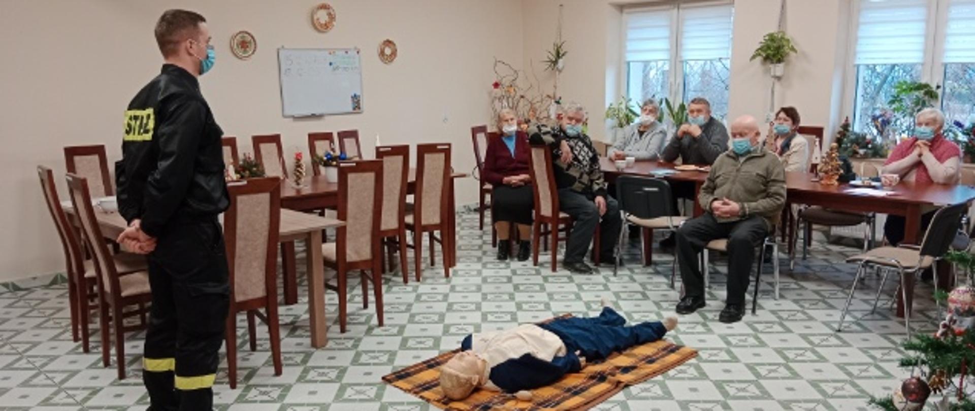 Umundurowany strażak w sali szkoleniowej stoi przed przed ułożonym na podłodze manekinem, w tle grupa starszych osób siedzących na krzesłach.