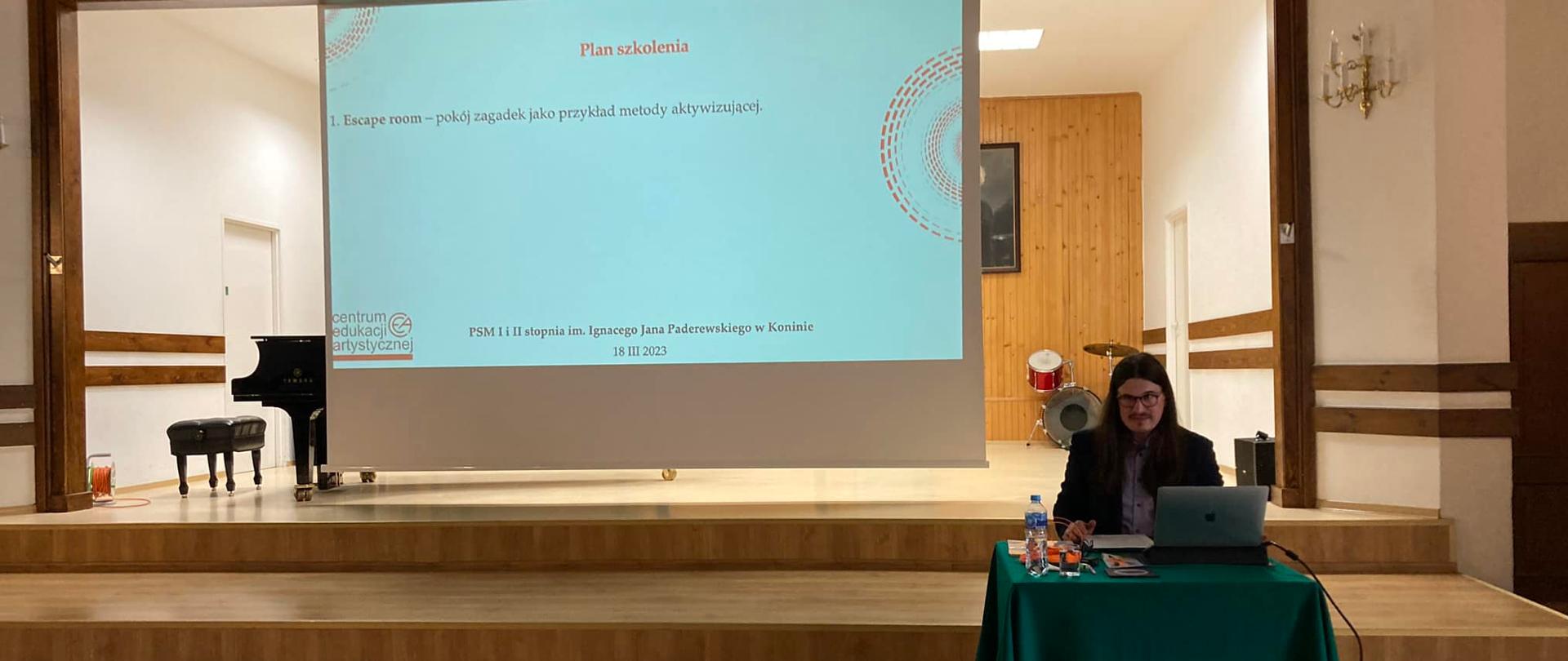 Prowadzący warsztaty siedzi na scenie PSM w Koninie, przy laptopie, z tyłu widać ekran z planem szkolenia.