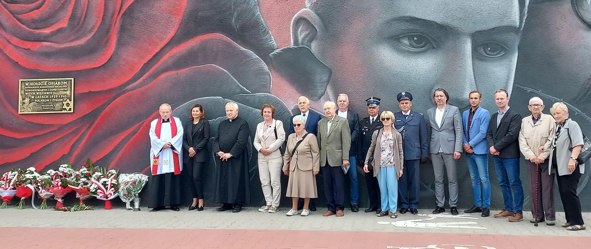 Narodowy Dzień Pamięci Ofiar Niemieckich Nazistowskich Obozów Koncentracyjnych i Obozów Zagłady. Na tle muralu znajdującego się na murze ZK w Rawiczu stoją uczestnicy uroczystości. Z lewej strony ksiądz. Obok tablica pamiątkowa, pod którą są wiązanki kwiatów.
