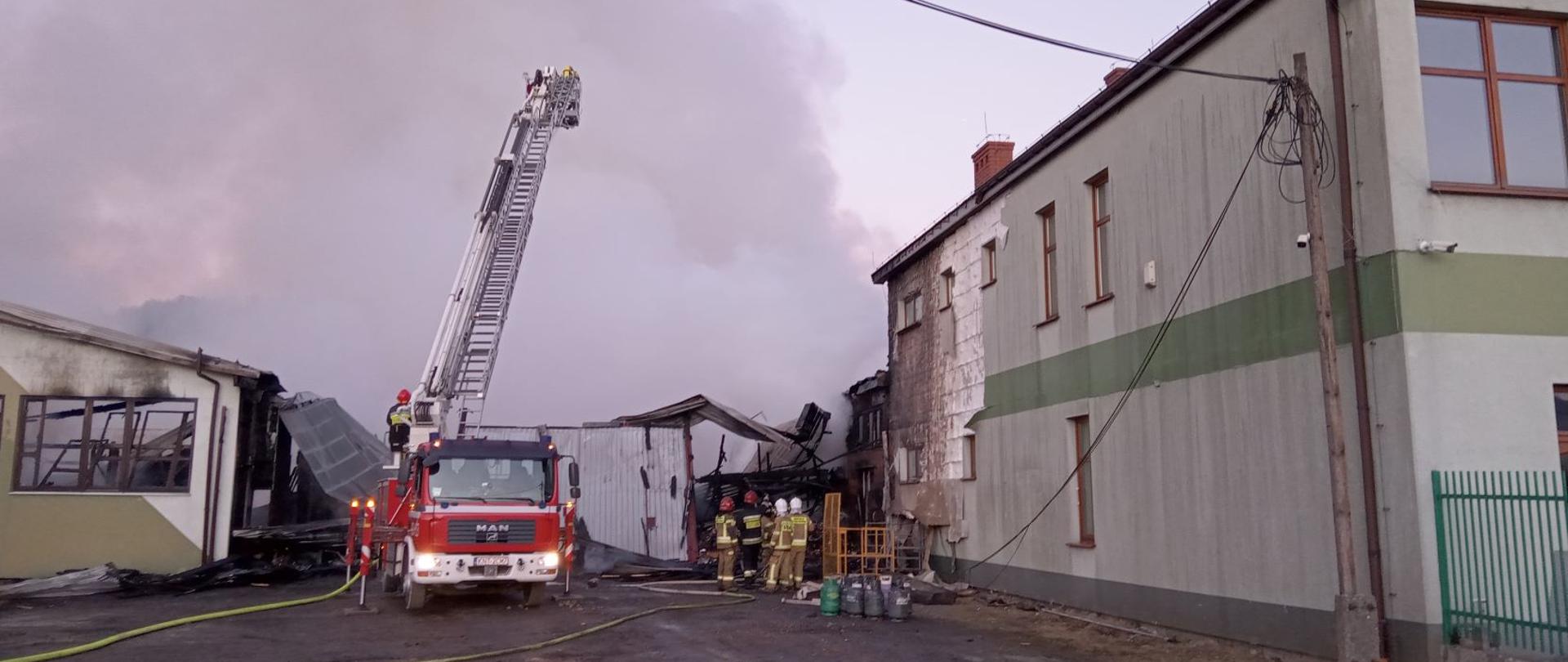 Zdjęcie przedstawia działania gaśnicze prowadzone od strony zachodniej. Widoczny podnośnik strażacki w którego koszu znajdują się strażacy. Z prawej strony grupa strażaków analizuje sytuację po wyciągnięciu z wnętrza butli gazowych 11 kg, oraz budynek administracyjno - biurowy, który udało się obronić.
