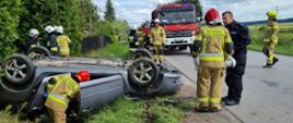 Na zdjęciu widać uszkodzony samochód osobowy znajdujący się na dachu w przydrożnym rowie. Obok samochodu znajduje się kilku strażaków w umundurowaniu specjalnym oraz policjant. W tle zaparkowane na poboczu drogi samochodu ratowniczo-gaśnicze.