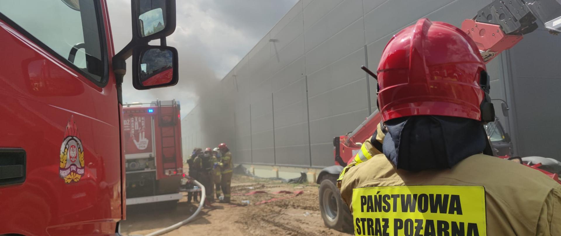 
Poziome, plenerowe ujęcie z poziomu ziemi, w słoneczny, częściowo pochmurny dzień, przedstawia duży, czerwony wóz strażacki zaparkowany na piaszczystym terenie obok ciemnoszarego budynku przemysłowego. Wóz strażacki, zwrócony lekko w prawo, ma biały przedni zderzak i tablicę rejestracyjną "PO 3WF97". Widać niebieskie światła alarmowe na dachu kabiny. Z tyłu wozu biegną czerwone węże strażackie. Po lewej stronie wozu strażackiego stoi strażak w żółtym kombinezonie i hełmie, odwrócony plecami do widza. W oddali, za pierwszym wozem, widać drugi czerwony wóz strażacki z numerem "469-21" na boku. Po prawej stronie zdjęcia leżą betonowe kręgi. W tle widać wiejskie domy z czerwonymi dachami i zieloną roślinność, a nad nimi częściowo zachmurzone niebo. Zgodnie z informacjami prasowymi, jest to pożar hali produkcyjno-magazynowej w miejscowości Gola, w powiecie kępińskim.