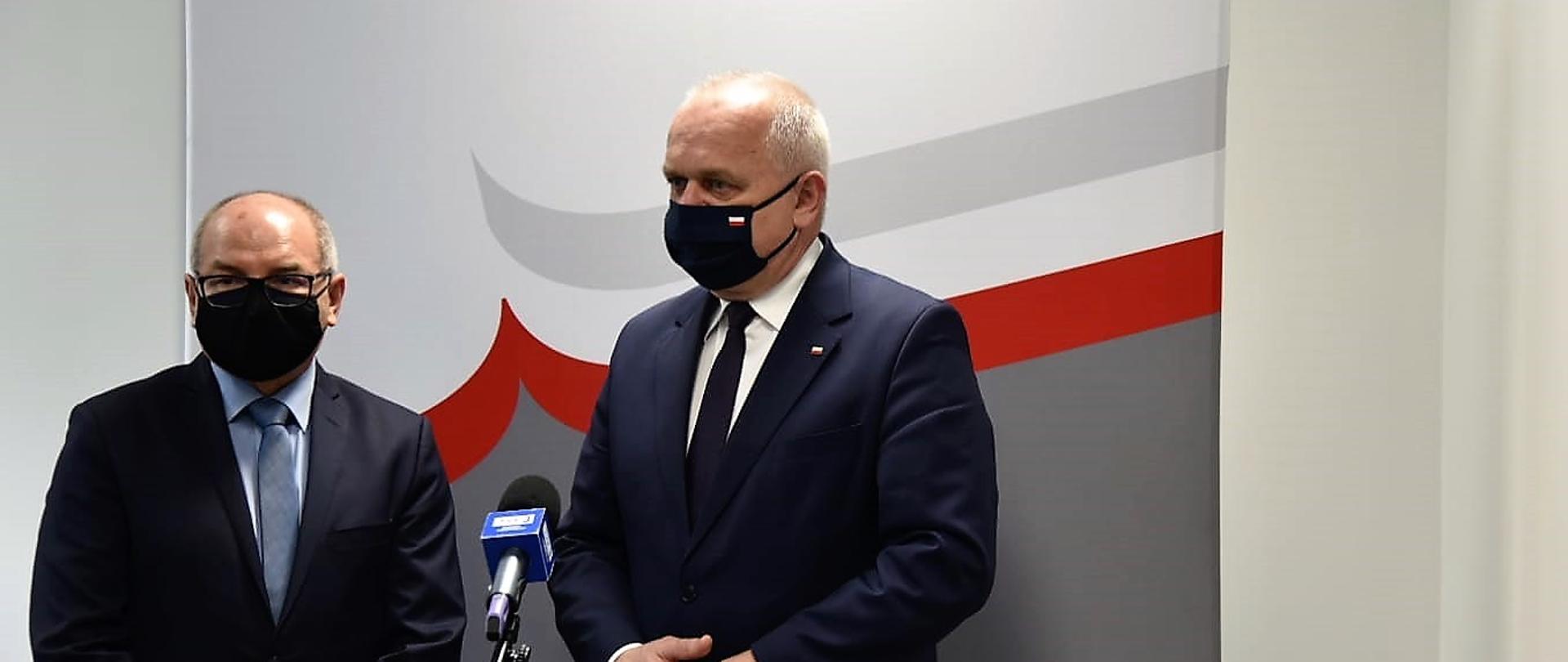 Wojewoda stoi z dyrektorem Wojewódzkiego Urzędu Statystycznego przed mikrofonem. Tuż za nimi ścianka promocyjna urzędu w kolorach biało-czerowno-szarych z logiem i godłem na górze 