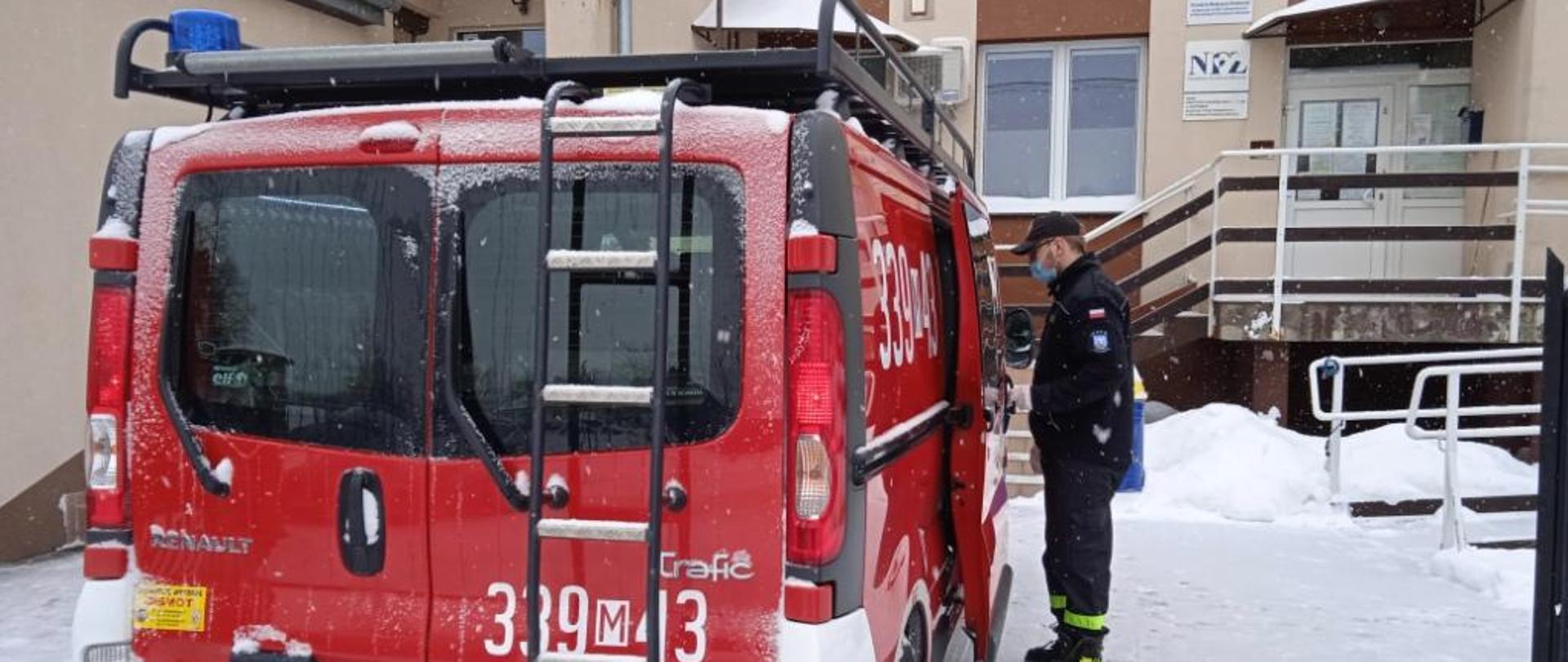 Na zdjęciu widać pojazd specjalny bus służący do przewozu osób, należący do Ochotniczej Straży Pożarnej w Jastrzębi. Pojazd zaparkowany jest przed Poradnią Ochrony Zdrowia w Jastrzębi. Prawe tylne drzwi przedziału pasażerskiego busa są otwarte. Przodem do nich stoi strażak ochotnik ubrany w czarne ubranie. Teren dookoła pokrywa śnieg. 