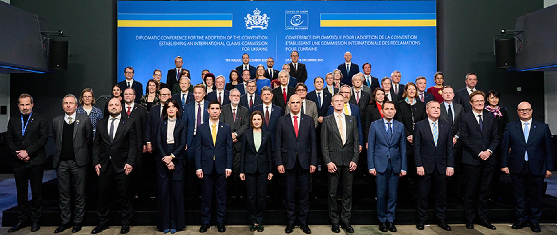 Na zdjęciu jest grupa kilkunastu osób, które stoją w pięciu rzędach. Są zwrócone w kierunku obiektywu. Mają na sobie ciemne, eleganckie ubrania. Za nimi jest niebieska ścianka. 