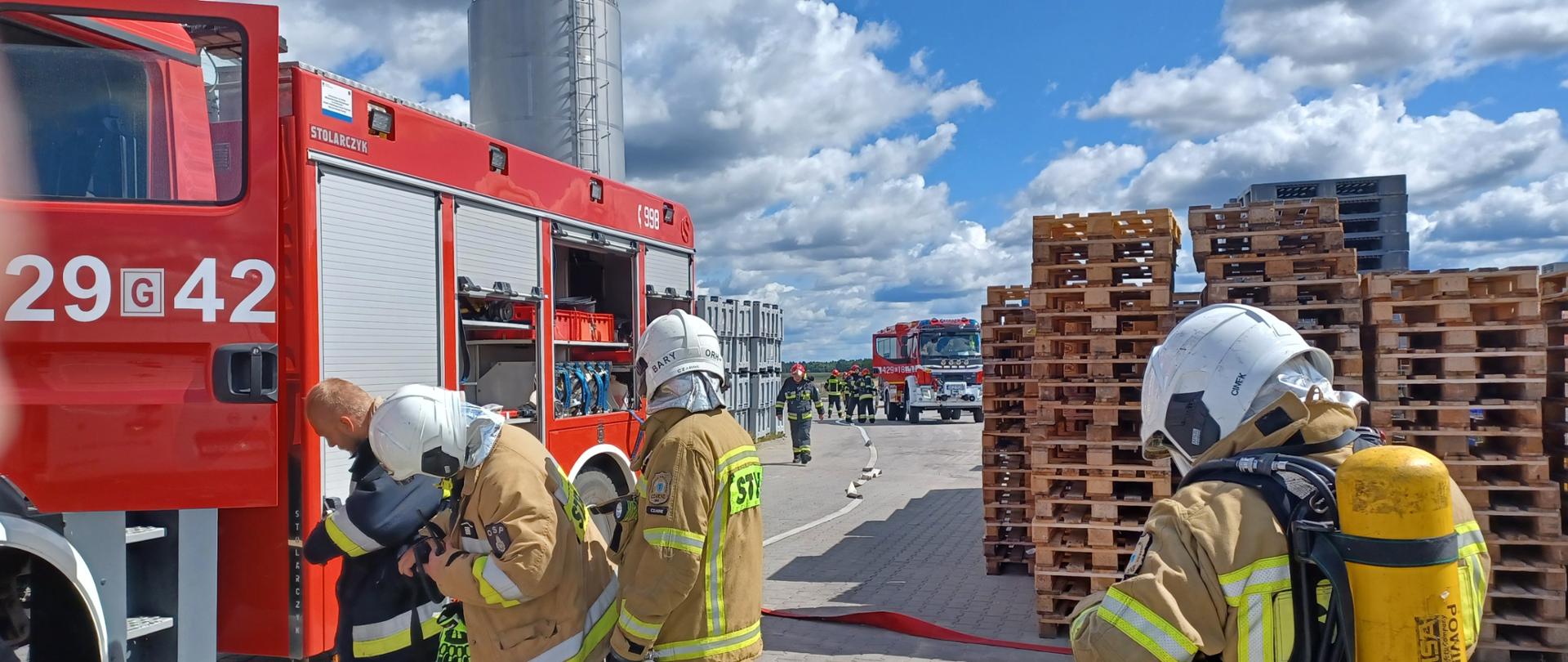 Na pierwszym planie widać strażaków zakładających sprzęt ochrony układu oddechowego i rozwijających węże pożarnicze. W tle stoją samochody pożarnicze.