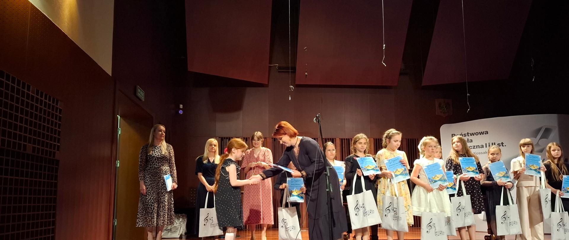 Uroczyste wręczenie dyplomów i upominków dzieciom na scenie szkoły muzycznej. Kobieta wręcza dyplom dziewczynce, podczas gdy inne dzieci stoją w rzędzie, trzymając podarki.