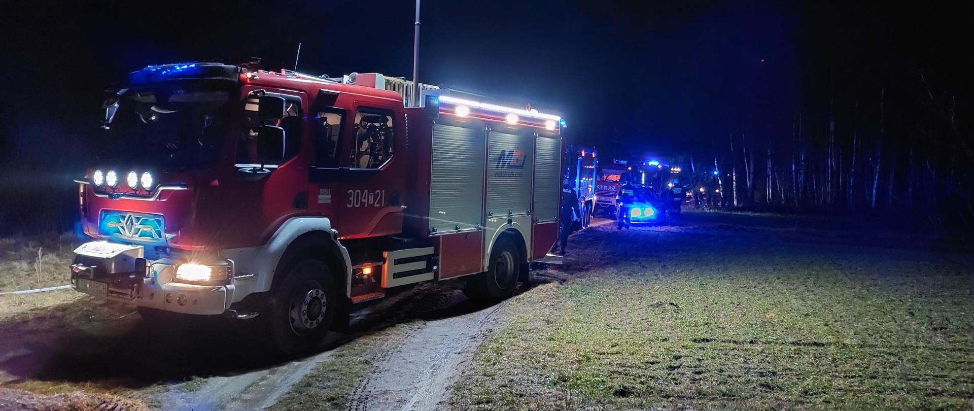  Zdjęcie przedstawia samochód pożarniczy stojący na drodze gruntowej skąd prowadzone są działania w oddalonym o około 150 m budynku mieszkalnym z powodu niemożności dojazdu. Pojazd ma wysunięty maszt oświetlający miejsce działań. W dali widać inne samochody strażackie.