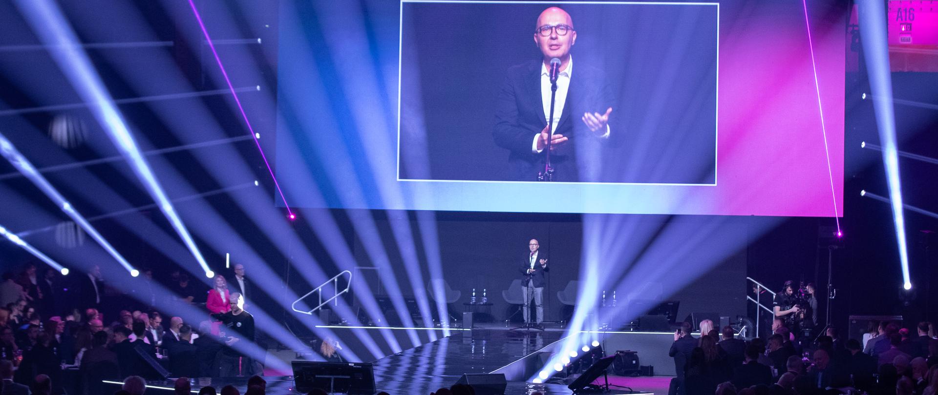 Wiceminister Robert Kropiwnicki przemawia ze sceny. U góry ekran. Ujęcie zza słuchaczy. 