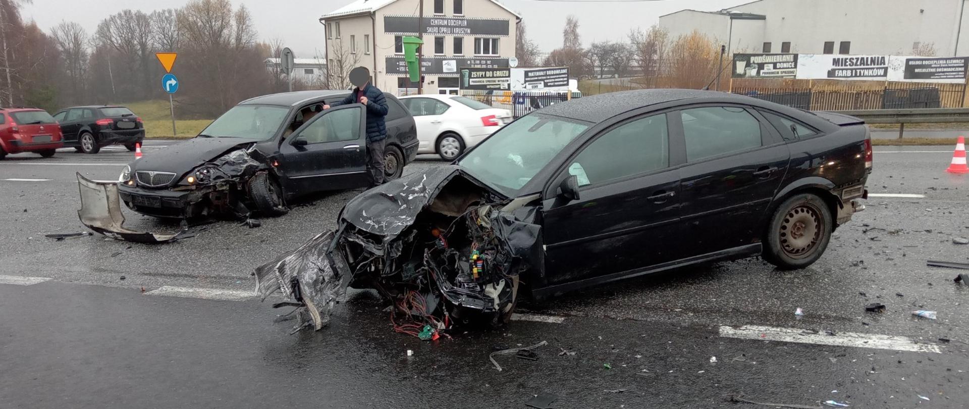 Zdjęcie przedstawia samochody osobowe zniszczone w wyniku zderzenia z innymi pojazdami znajdujące się na jezdni drogi krajowej nr 7. W tle widoczne zabudowania miejscowości i pojazdy przejeżdżające otwartym pasem ruchu w kierunku Krakowa.