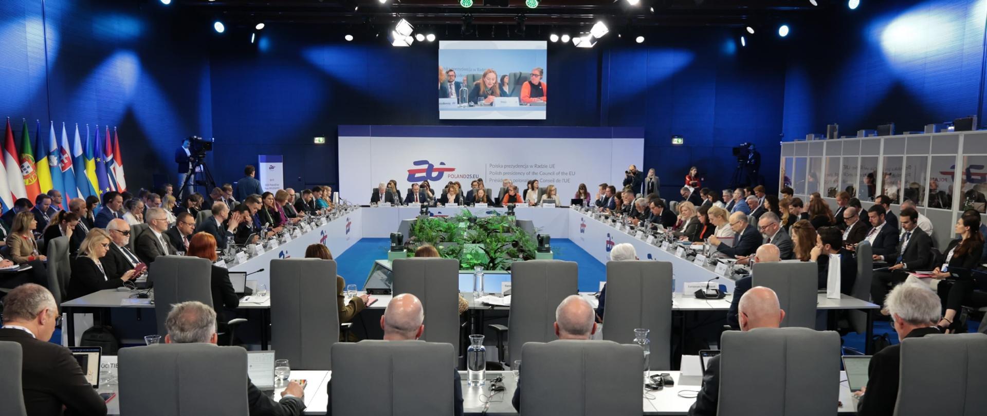 W sali konferencyjnej przy stołach siedzą osoby. Na górze na ekranie minister Katarzyna Pełczyńska-Nałęcz. Przy ścianie flagi.