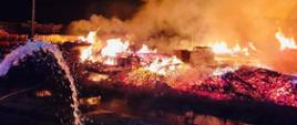 Zdjęcie przedstawia pożar hali produkcyjnej oraz magazynowej w miejscowości Muchy. Na zdjęciu widzimy rozwinięty pożar magazynów palet objęty całościowo ogniem. Po lewej stronie widoczny prąd wody gaszący pożar. Zdjęcie wykonane w porze nocnej na placu składowym w zakładzie.
