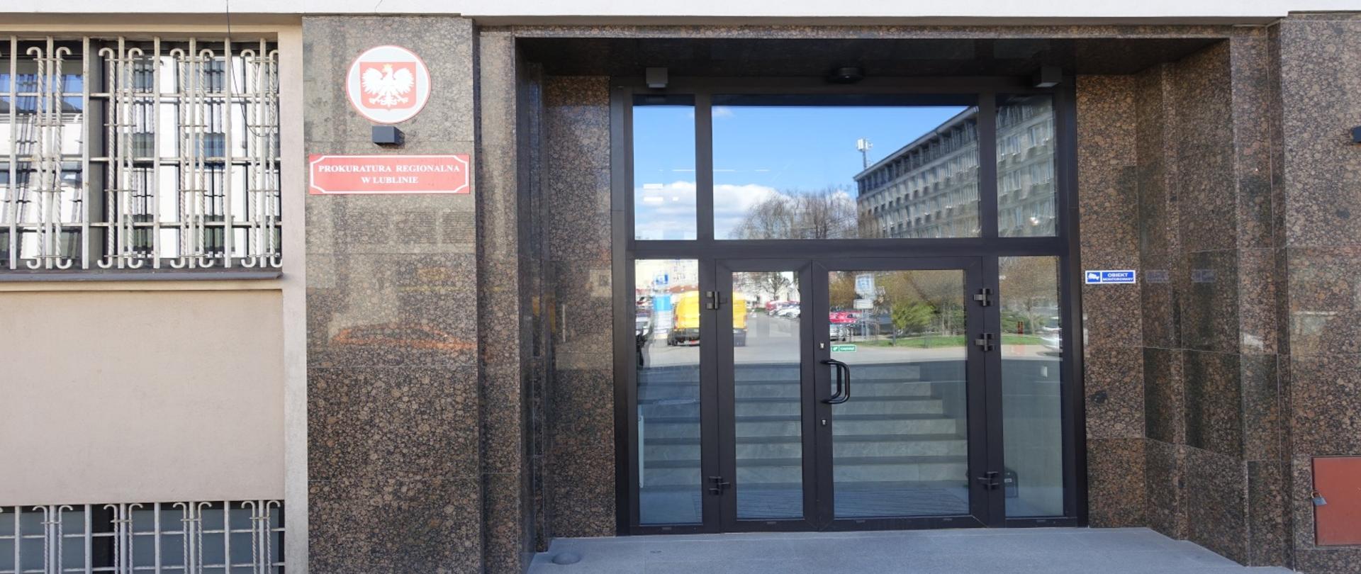 Wejście do budynku Prokuratury. Na ścianie godło oraz czerwona tabliczka z białymi napisami