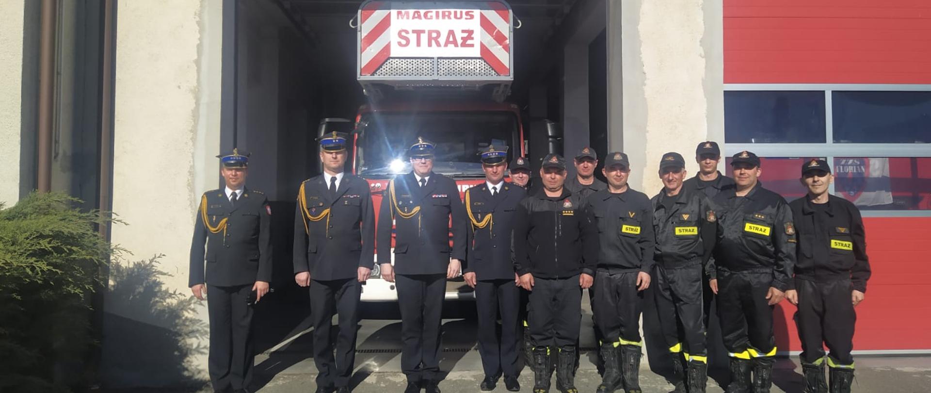 Na zdjęciu czterech funkcjonariuszy Państwowej Straży Pożarnej w umundurowaniu wyjściowym, oraz strażacy w strojach koszarowych ustawieni przed budynkiem Jednostki Ratowniczo-Gaśniczej w Koniecpolu