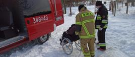 Strażacy wiozą kobietę na wózku inwalidzkim do samochodu strażackiego
