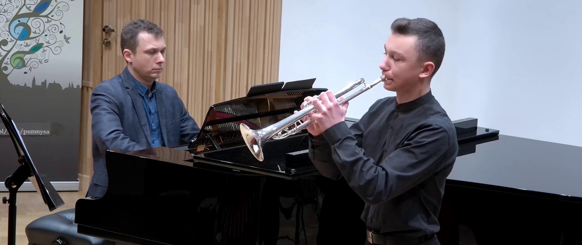 Grafika - zdjęcie. Na estradzie sali koncertowej występuje trębacz wraz z towarzyszącym mu pianistą. W tle z lewej strony fragment baneru PSM Nysa.