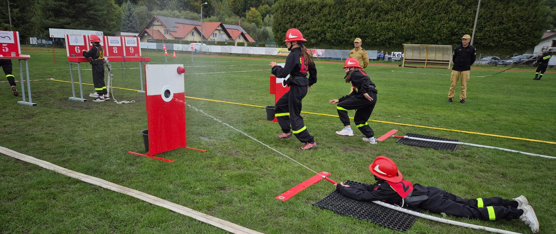 Na zdjęciu znajdują się zawodnicy startujący w konkurencji ćwiczenia bojowego, obok sędziowie. Na pierwszym planie zawodnik leżąc celuje z prądownicy wodnej do nalewaka. W dalszym planie pozostali zawodnicy wykonujący zadania. 