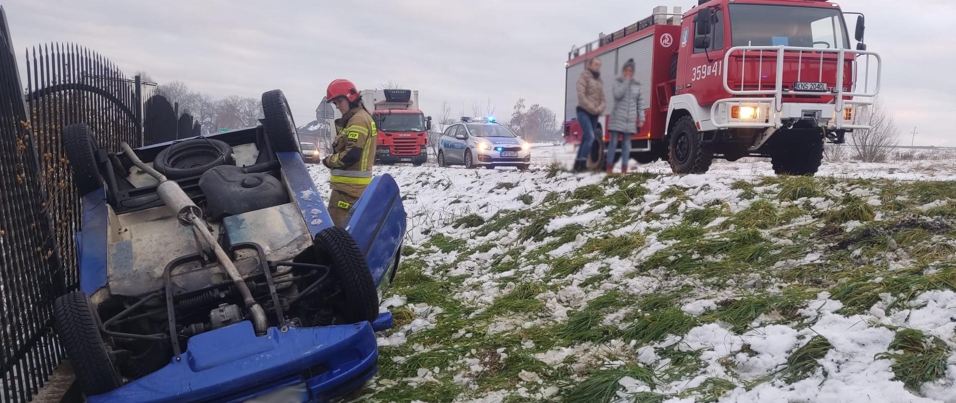 Samochód osobowy poza drogą na dachu obok ogrodzenia. Na drodze samochód pożarniczy.