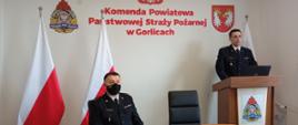 Na zdjęciu widoczny po prawej stronie na tle białej ściany z godłem Rzeczpospolitej Polskiej napisem w kolorze czerwonym Komenda Powiatowa Państwowej Straży Pożarnej w Gorlicach po bokach flagi biało czerwone przemawiający z-ca komendanta powiatowego Państwowej Straży Pożarnej w Gorlicach st.kpt. Waldemar Krok po lewej stronie siedzący komendant powiatowy Państwowej Straży Pożarnej bryg. Krzysztof Gładysz organizujący roczną odprawę.
