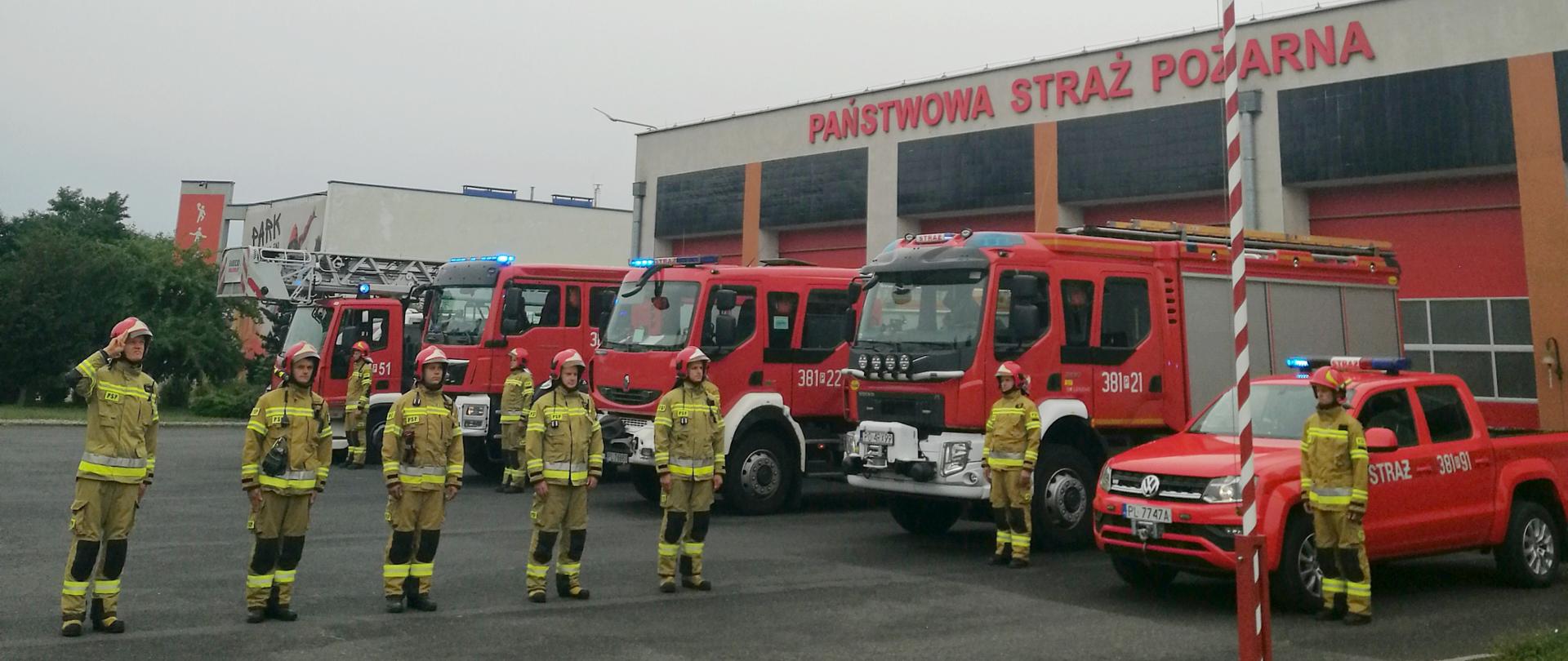 Strażacy stoją w szeregu przy maszcie flagowym. W tle widoczne samochody pożarnicze oraz garaże komendy.