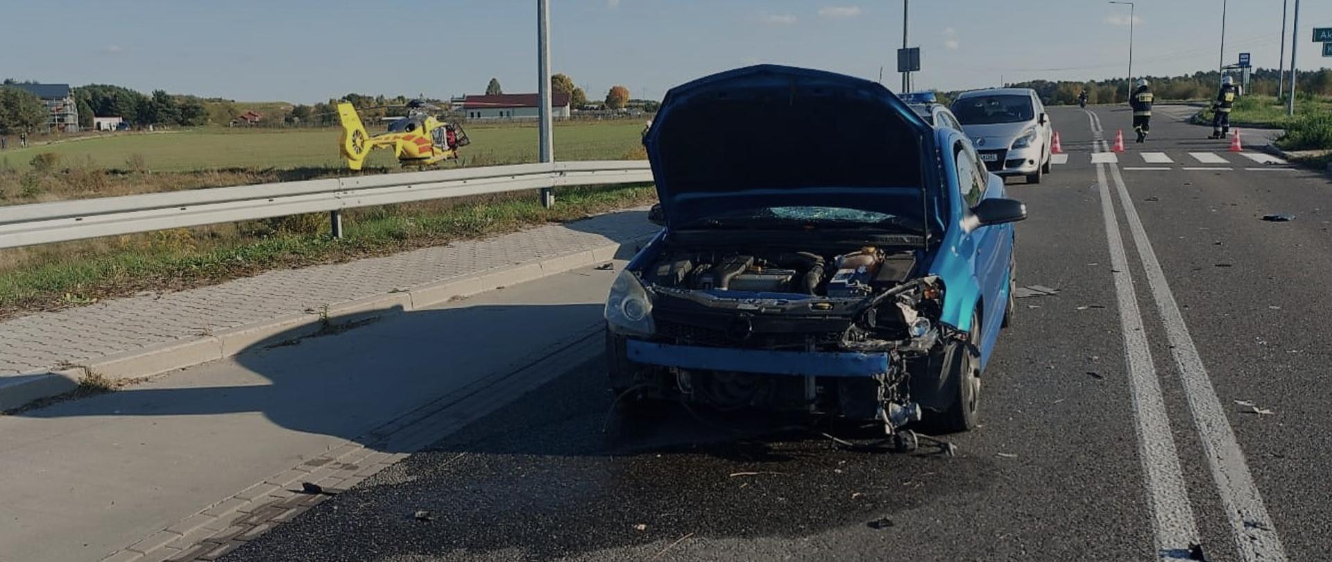 Zdjęcie przedstawia drogę wojewódzką na której doszło do wypadku komunikacyjnego. Na pasie drogowym stoi uszkodzony samochód osobowy. Po lewej stronie, na pobliskiej łące wylądował śmigłowiec LPR-u. Za autem, po prawej stronie widać dwóch strażaków.