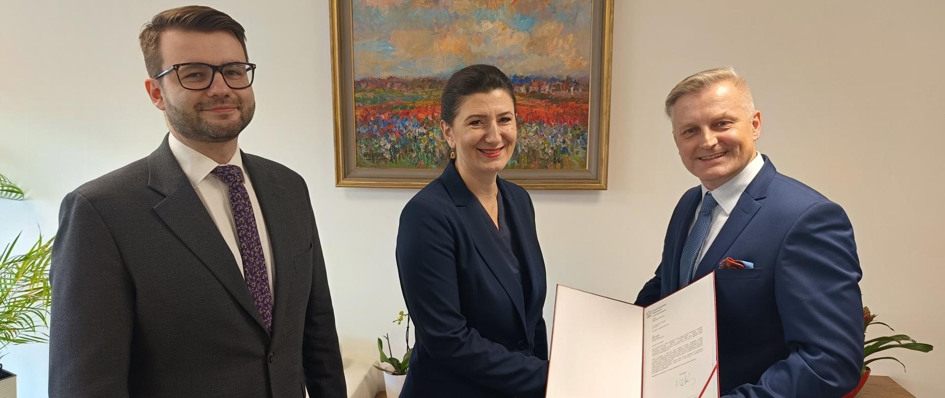 Zdjęcie trzech osób. W tle obraz z pejzażem wiszący na ścianie. Na zdjęciu od lewej: Pan Przemysław Grosfeld, Wiceprezes UZP, Pani Agnieszka Olszewska, Prezes UZP oraz Pan Rafał Opaliński, Prezes Fundacji Dzieci-Dzieciom