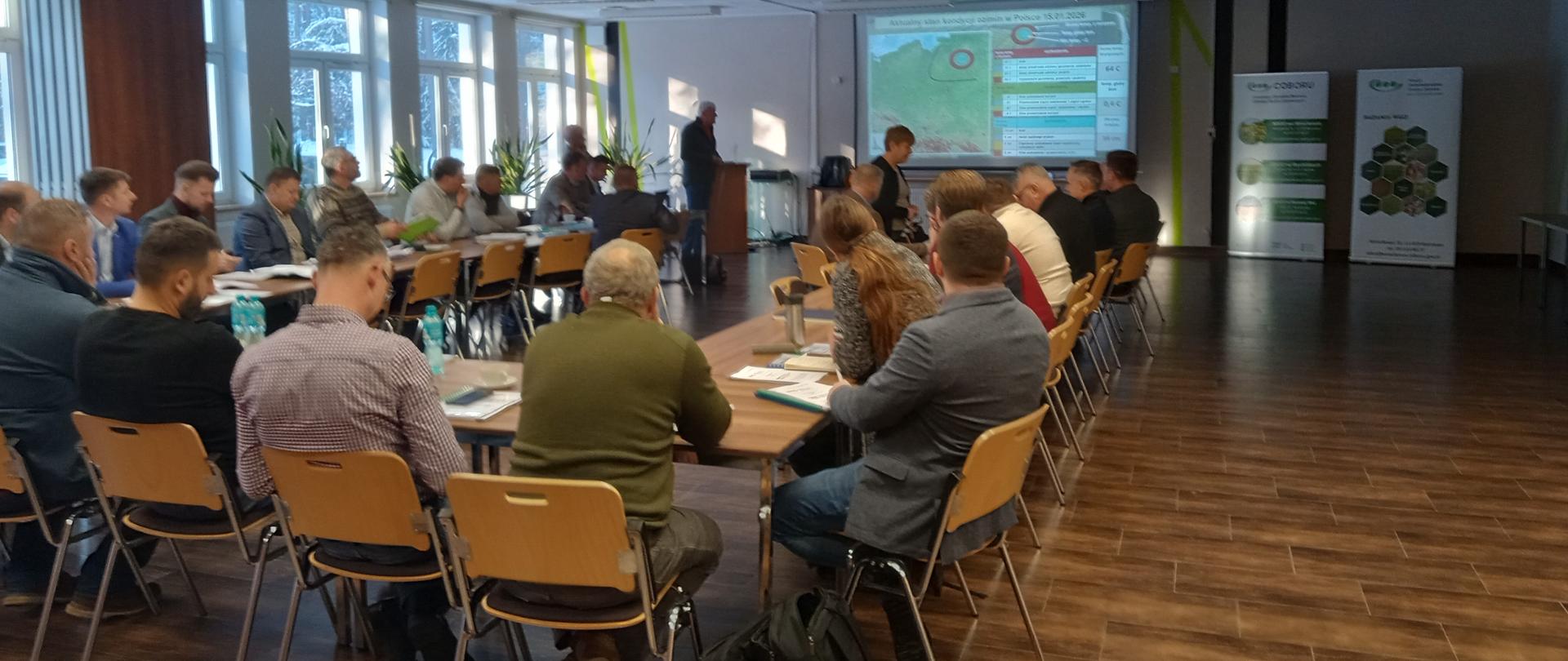 grupa osób siedzi przy stołach, w głębi prezentacja