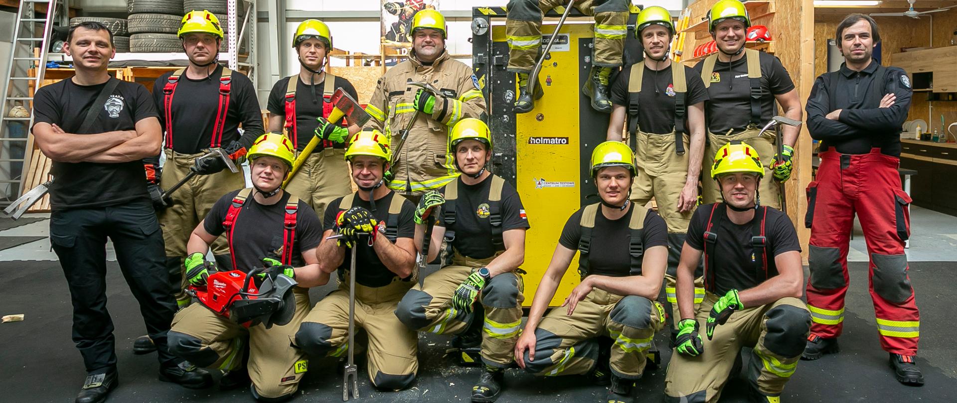 Zdjęcie grupowe strażaków. Ratownicy stoją w dwóch rzędach, w rękach trzymają podręczny sprzęt burzący. W centralnej części znajduje się trenażer do siłowego otwierania drzwi.