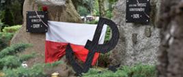 Na kamiennych blokach przykręcone czarne tabliczki z napisami "gen. dyw. Stefan Rowecki GROT" i "gen. dyw. Tadeusz Komorowski BÓR". Za tablice zatrknięte pojedyncze kwiaty. Pomiędzy kamieniami rozwieszona biało-czerowna znak Powstania z literą P wpisaną w kotwice.