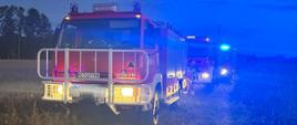 Wóz strażacki z włączonymi światłami stoi na polnej drodze, za nim kolejny pojazd gaśniczy. W tle zmierzch i zachmurzone niebo.