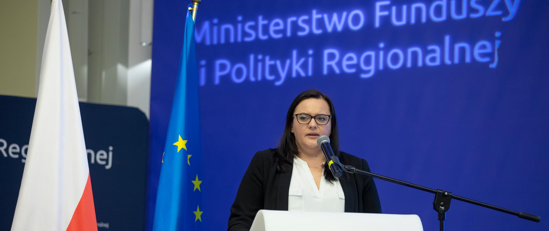 Wiceminister Małgorzata Jarosińska-Jedynak stoi w białej mównicy z mikrofonem. Obok kwiaty w donicy i dwie flagi PL i UE. Z tyłu ścianka z napisem Ministerstwo Funduszy i Polityki Regionalnej.