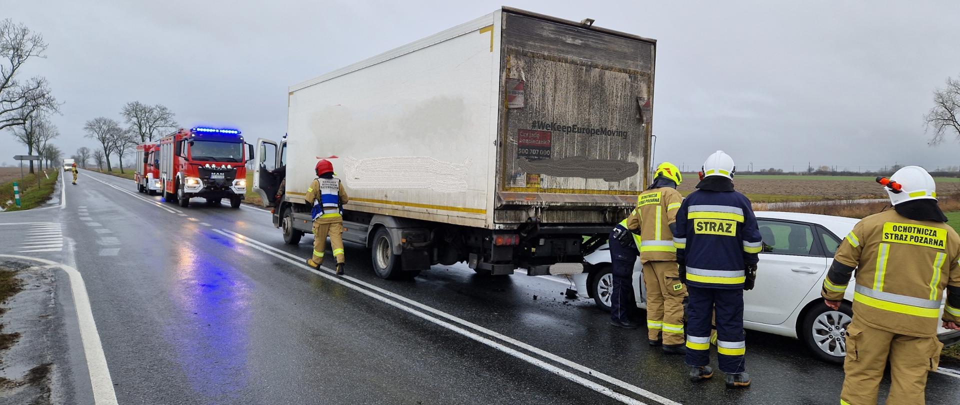 Zdjęcie przedstawiają samochody biorące udział w zdarzeniu oraz pojazdy straży pożarnej, które stoją na jednym pasie drogi. Przy pojazdach stoją strażacy wykonujący czynności służbowe.