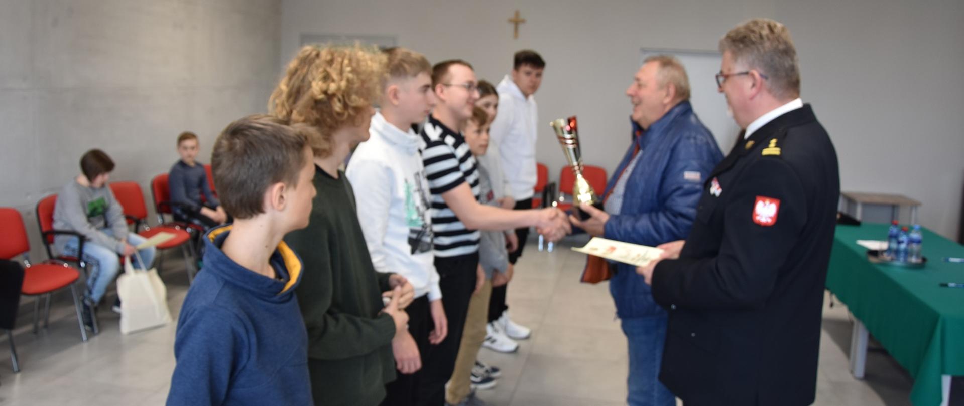Zdjęcie obrazuje młodzież strażak i osobę wręczającą uczniowi puchar
