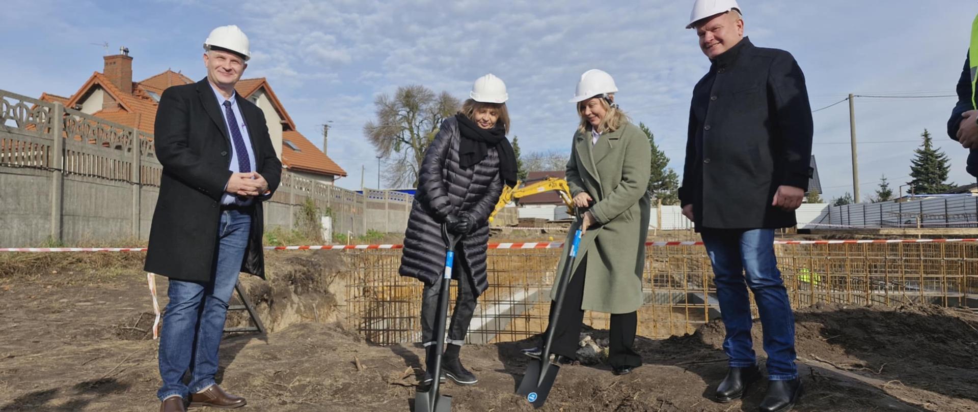 Plac budowy, dwie kobiety wkopują łopaty w grunt. Obok stoi dwóch mężczyzn. Wszyscy mają na głowach kaski ochronne. W tle prace budowlane.