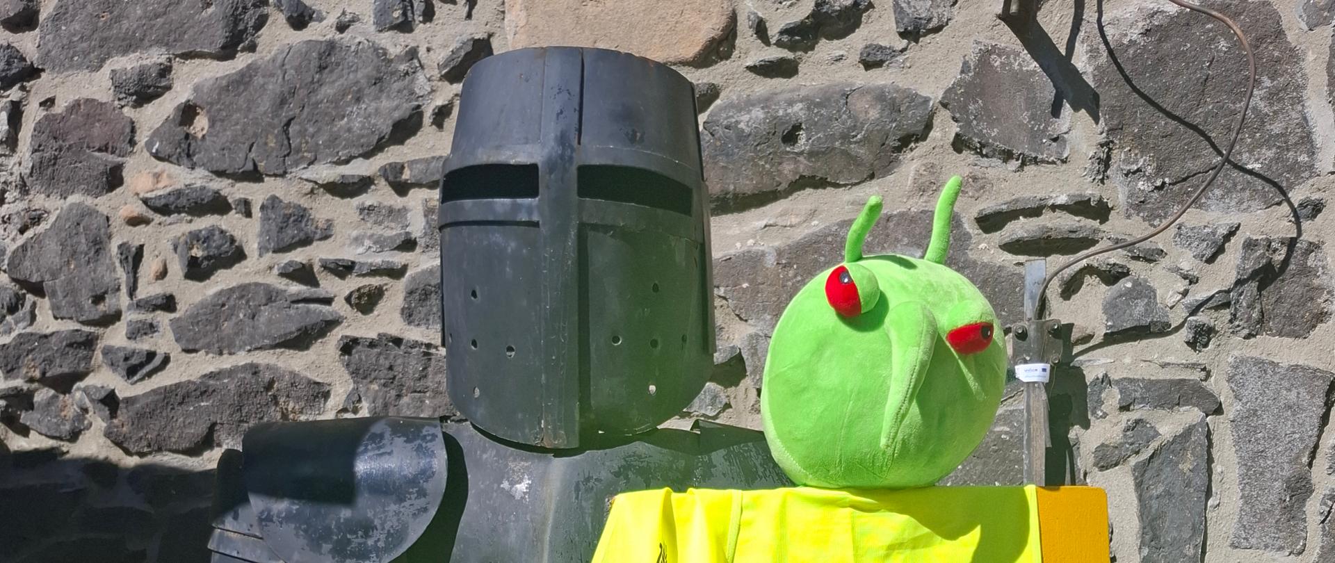 Maskotka Pesty siedząca na tarczy rycerza w zbroi. Tarcza jest przysłonięta zieloną kamizelką z napisem PIORiN