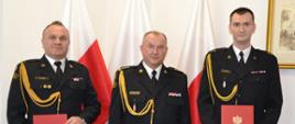 Trzech strażaków w mundurach wyjściowych ze sznurem stoją obok siebie skrajni strażacy trzymają czerwone teczki za nimi znajdują się trzy flagi Polski na ścianie wisi obraz. 