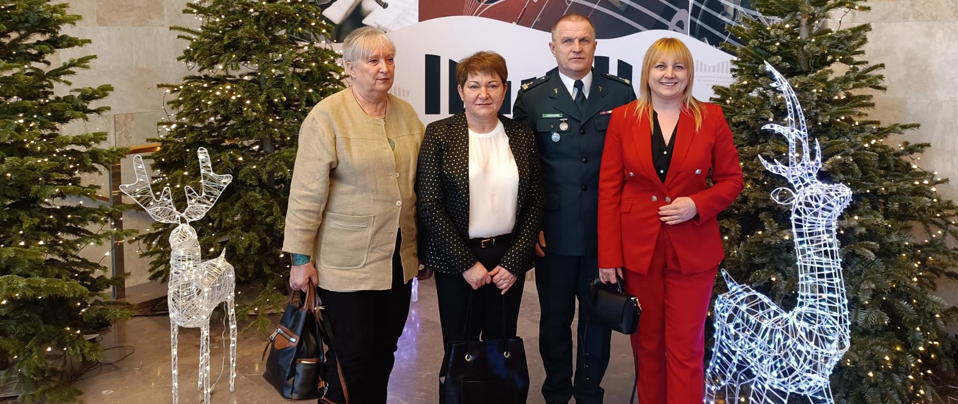 Trzy kobiety i jeden mężczyzna stoją w pomieszczeniu. Za nimi znajdują się choinki a obok nich stoją ozdobne świecące renifery. 