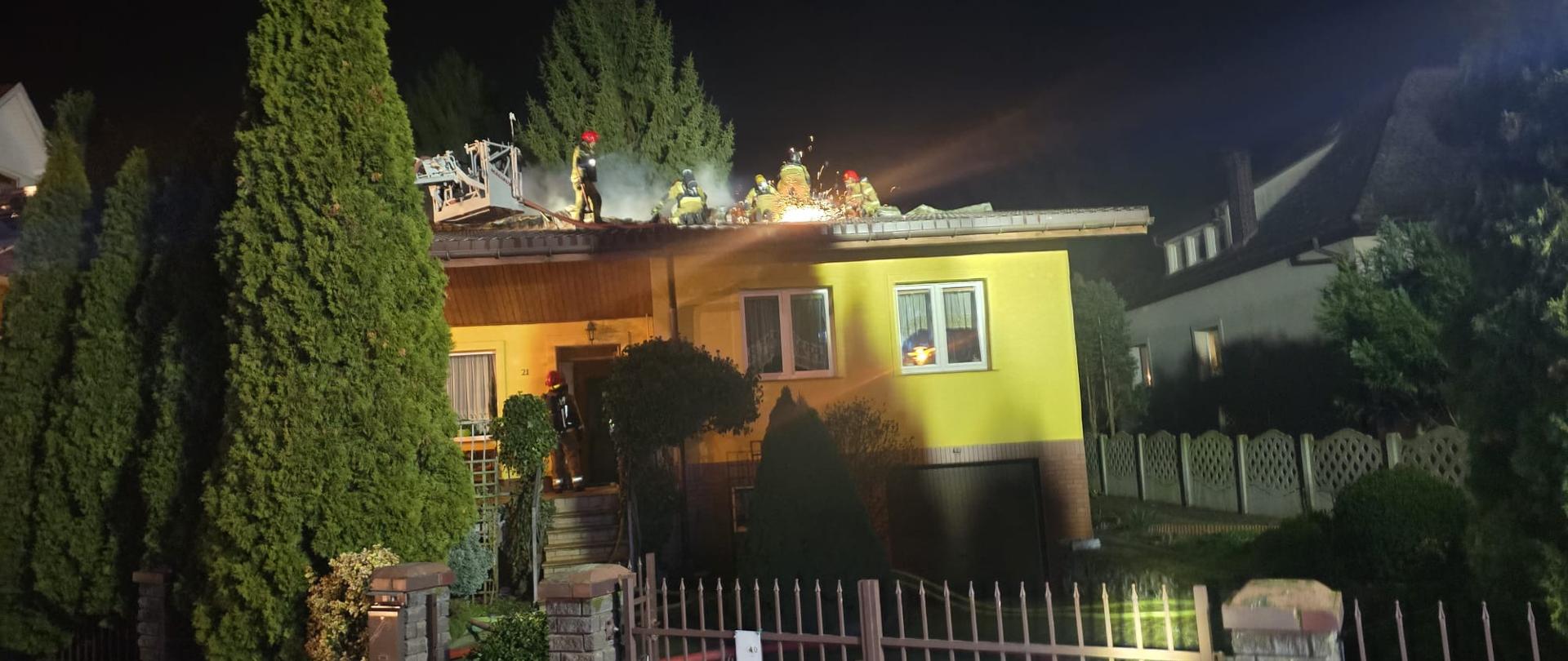 Na zdjęciu widać nocną akcję gaśniczą prowadzoną przez strażaków na dachu jednokondygnacyjnego domu jednorodzinnego w zabudowie jednorodzinnej. Strażacy w aparatach ODO pracują na dachu przy użyciu sprzętu burzącego i przecinającego, a z otworów w poszyciu wydobywa się dym; teren przed budynkiem jest oświetlony światłami pojazdów pożarniczych, a na pierwszym planie widoczne jest ogrodzenie posesji.