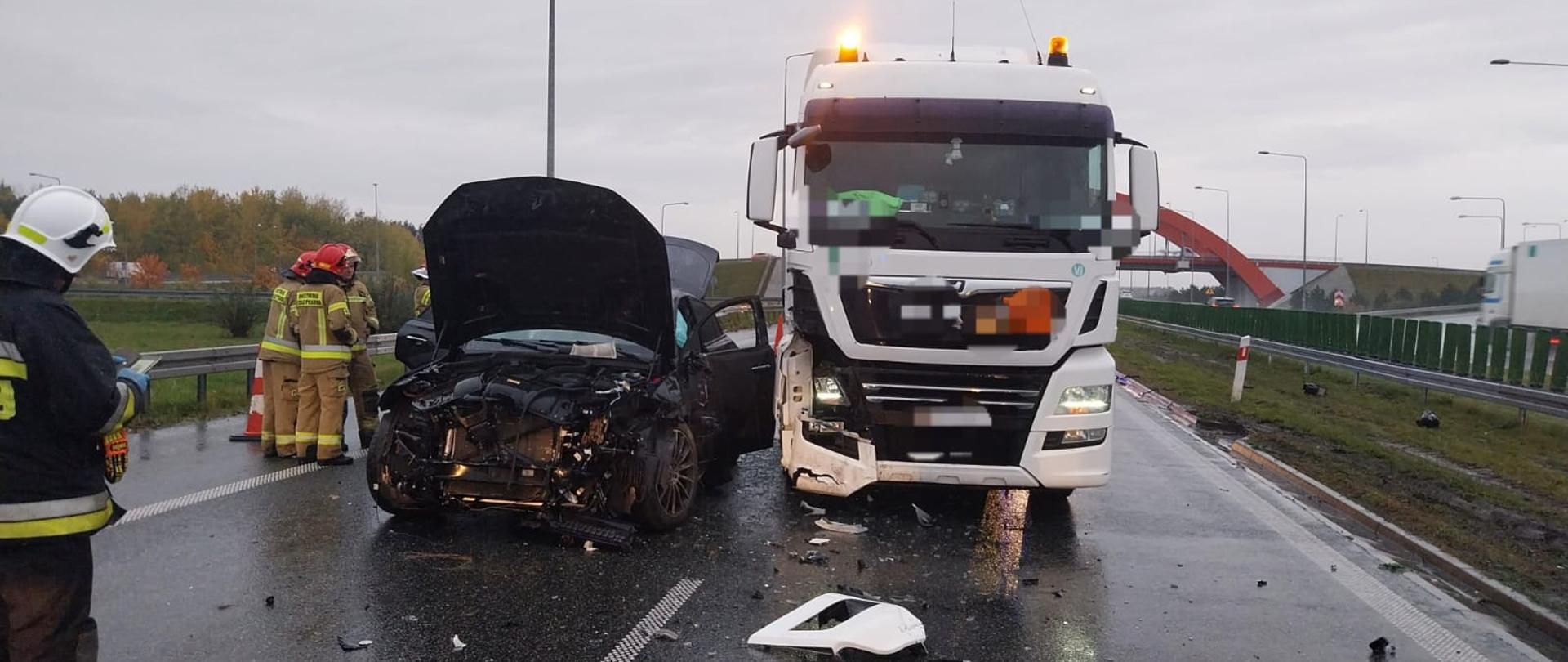 Na zdjęciu widać dwa rozbite pojazdy. Z lewej strony stoi czarny samochód osobowy z rozbitym przodem. Ma otwartą maskę oraz drzwi. Obok stoi biały samochód ciężarowy, który ma rozbity prawy przód. Oba pojazdy stoją na asfaltowej jezdni autostrady. Obok na jezdni stoją straży w żółtych strojach i czerwonych hełmach. Na pierwszym planie stoi jeden strażak w ciemnym stroju i białym hełmie. Na jezdni porozrzucane są elementy karoserii z pojazdów. W tle widać latarnie, drzewa oraz wiadukt. Jest widno, ale pochmurnie.