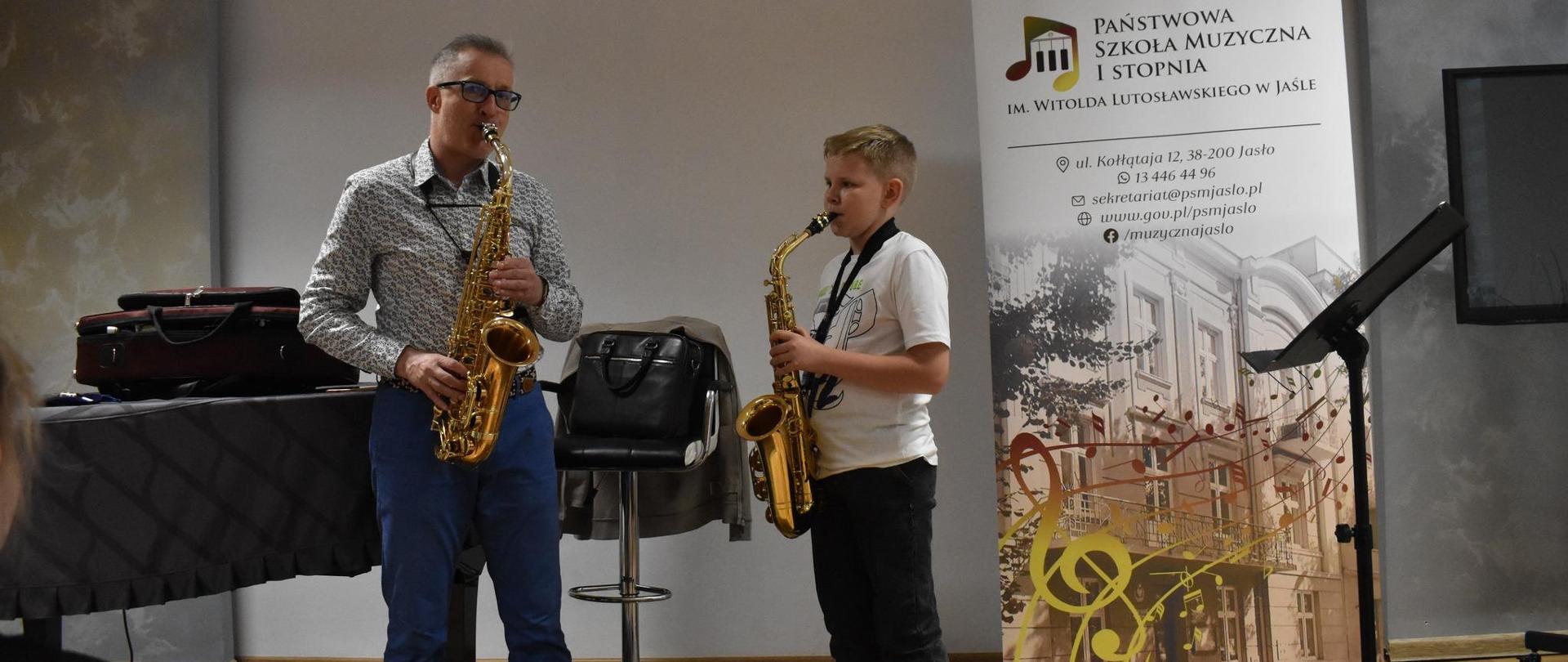 Pan saksofonista oraz uczeń stoją w Sali kameralnej PSM grając na saksofonie. Z prawej strony stoi czarny pulpit oraz rollup PSM. Ogólna kolorystyka szaro-brązowo-czarna.