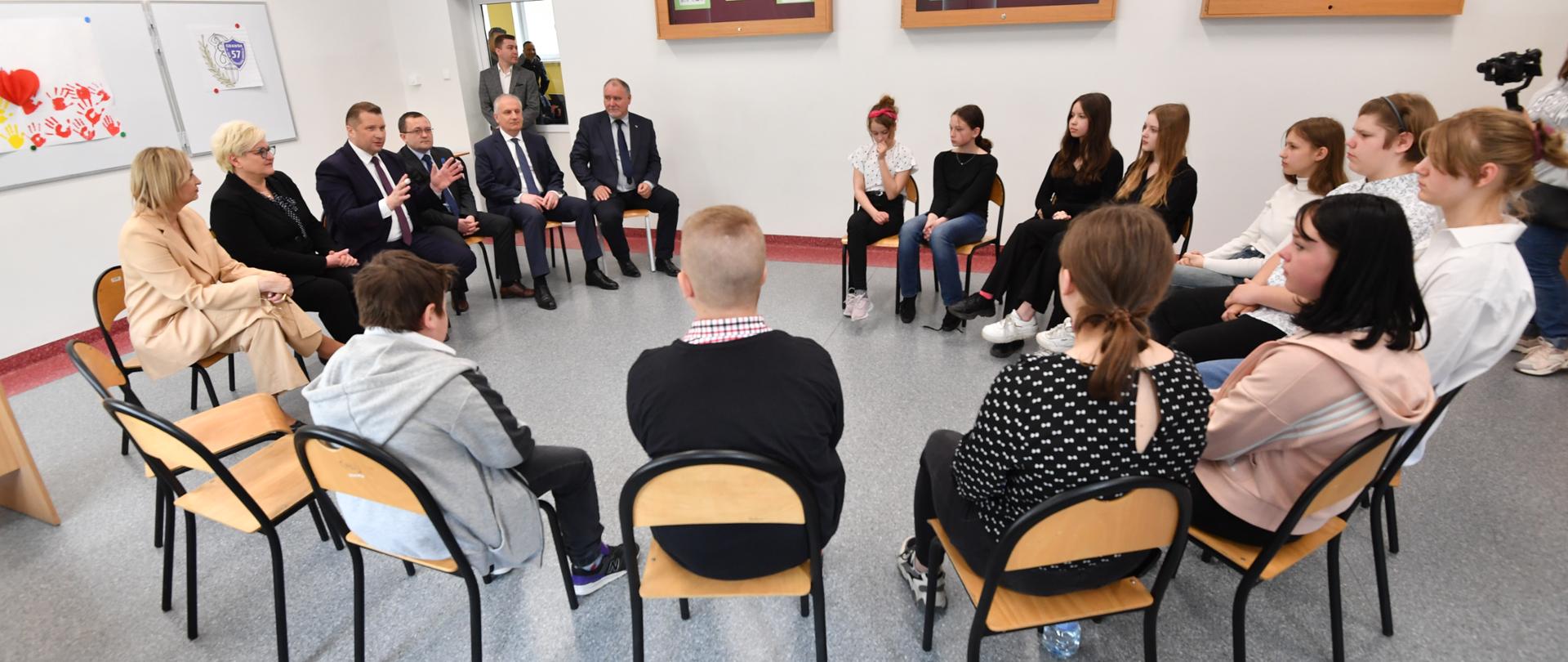 Klasa lekcyjna w krzesłami ustawionymi w okrąg. Krzesłach siedzą dzieci, nauczycielei i uczniowie oraz minister Czarnek, który gestykuluje coś rękoma. 