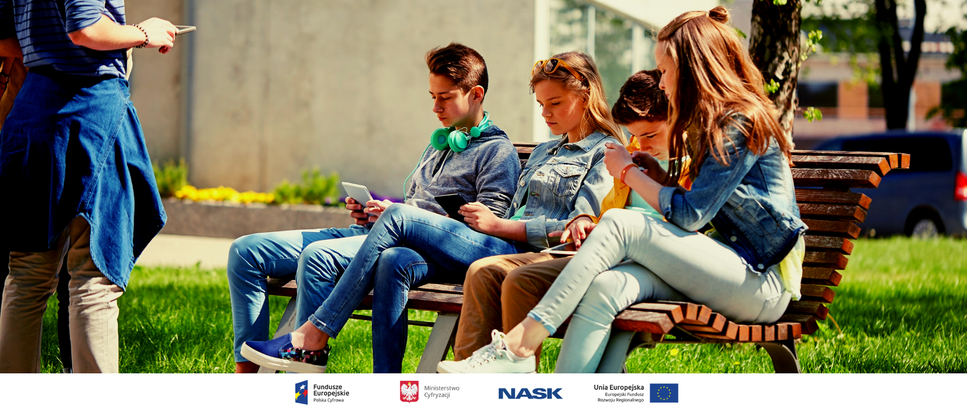 Na ławce siedzi czworo nastolatków patrzących w tablety i telefony. Obok stoi kolejna osoba z telefonem w ręce.