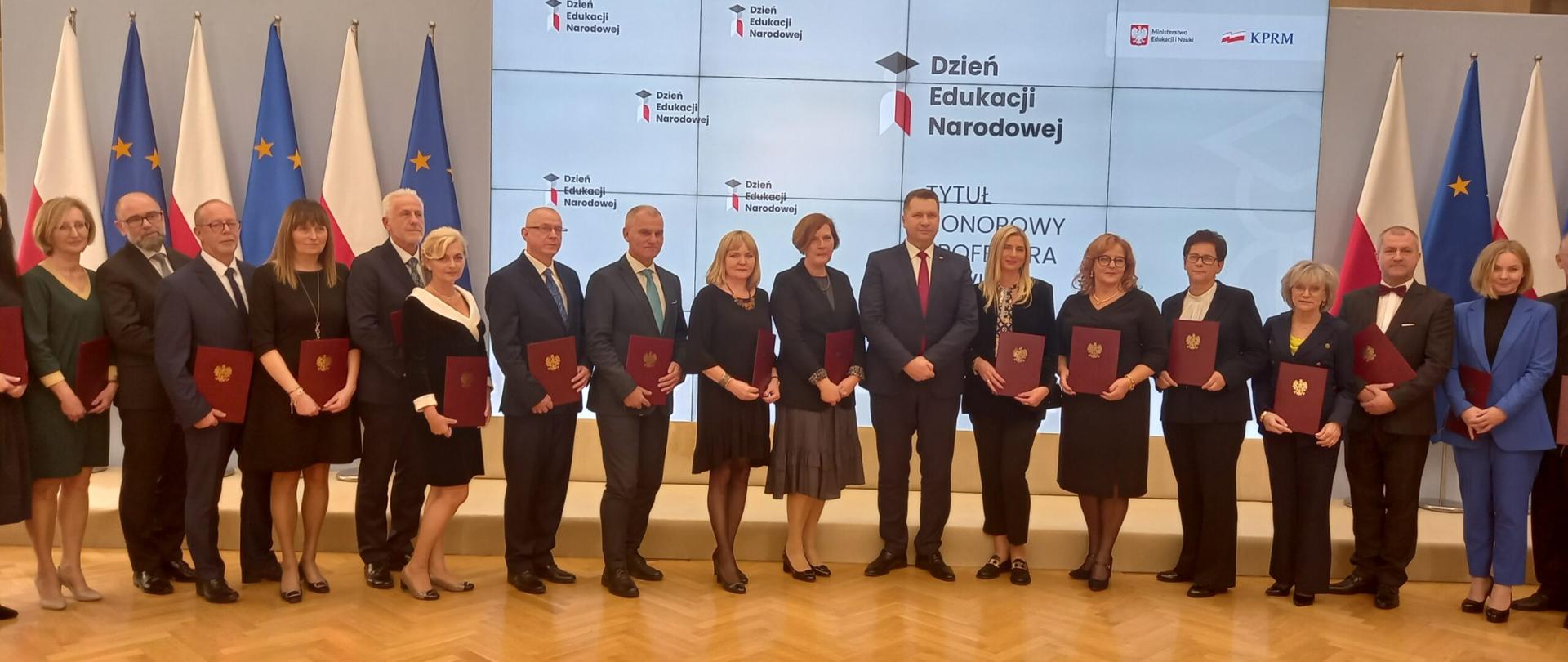 Grupowe zdjęcie nauczycieli, którzy otrzymali tytuł honorowego Profesora Oświaty, razem z Ministrem Edukacji Prof. Przemysławem Czarnkiem. W tle widać flagi Polski i Unii Europejskiej oraz napis: „Dzień Edukacji Narodowej ”.