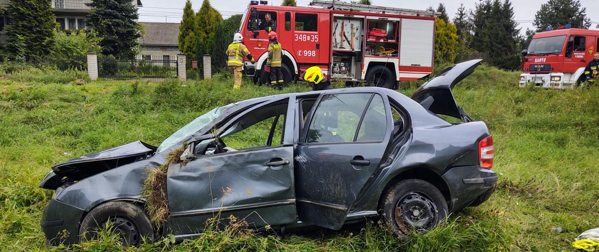 Samochód osobowy na łące z podniesioną klapą bagażnika. Na drodze samochody pożarnicze oraz strażacy.